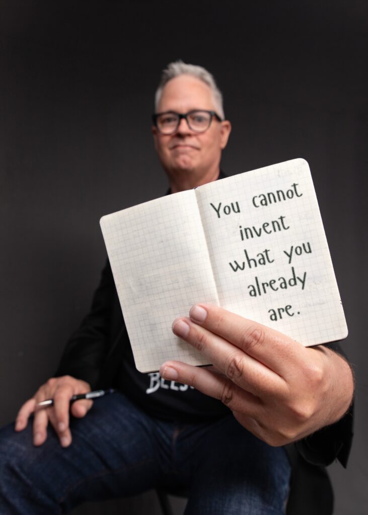 Tim Miles holding an open pocket notebook, on which is written "You cannot invent what you already are."