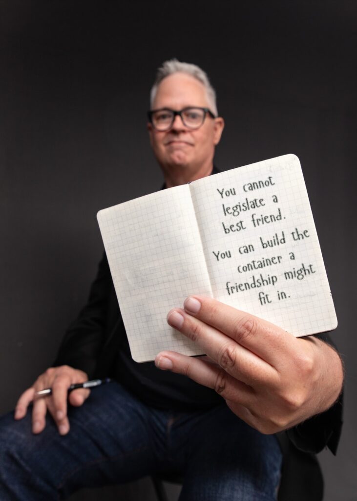 Tim Miles holding an open pocket notebook, on which is written "You cannot legislate a best friend. You can build the container a friendship might fit in."